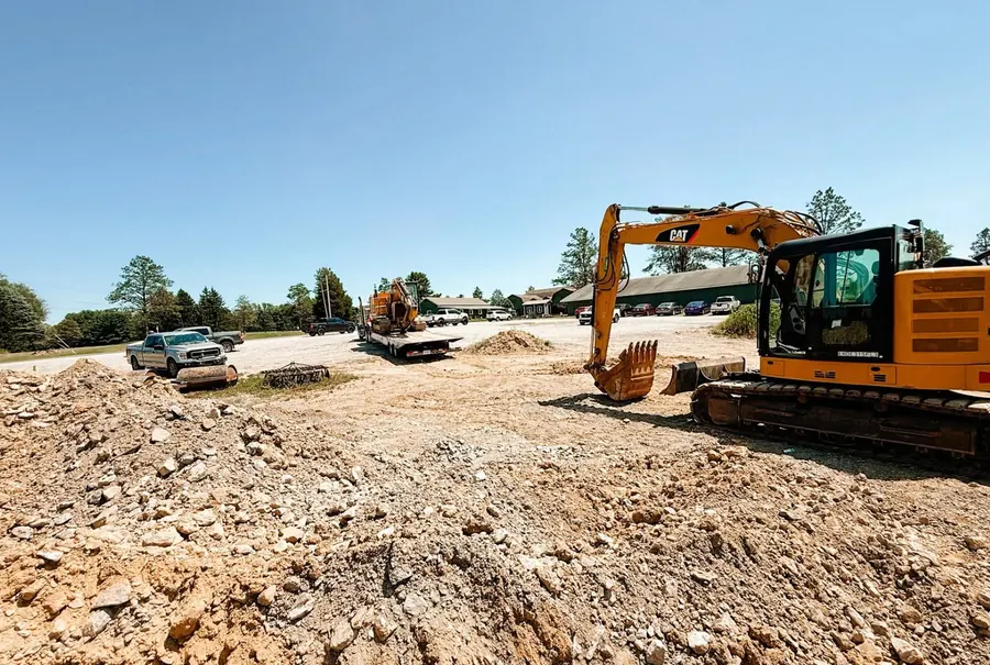 CAT excavator on a gravel lot with rock piles and a second machine on a trailer