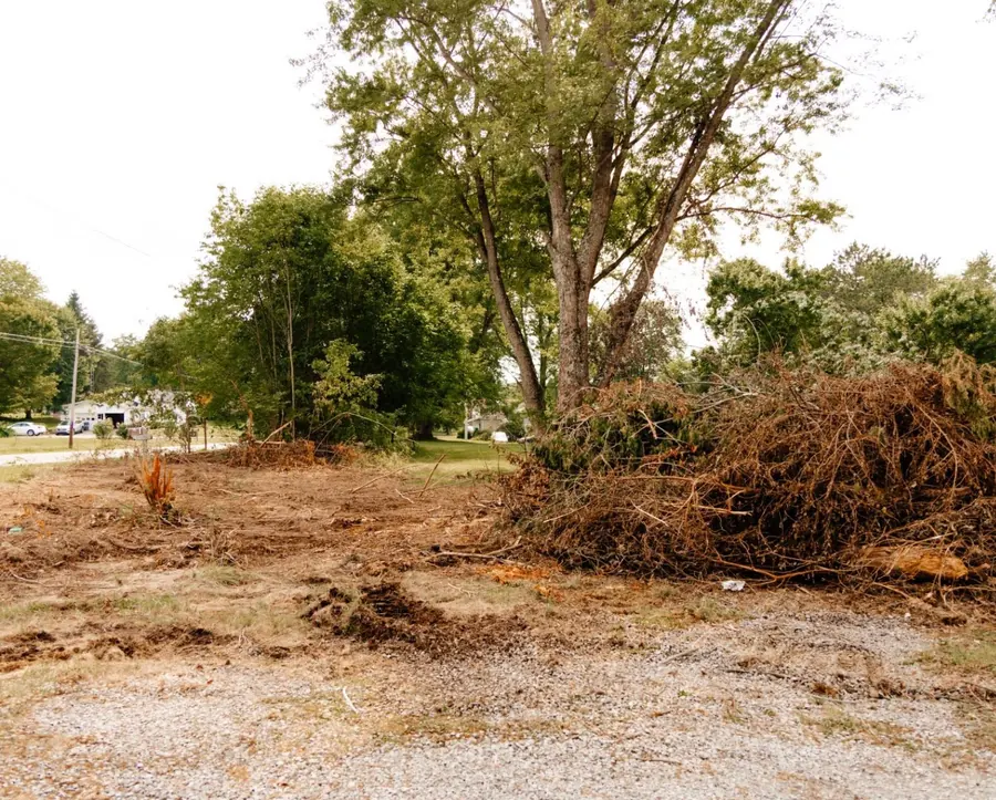 Cleared lot with a large brush pile beside a gravel pad and roadside trees