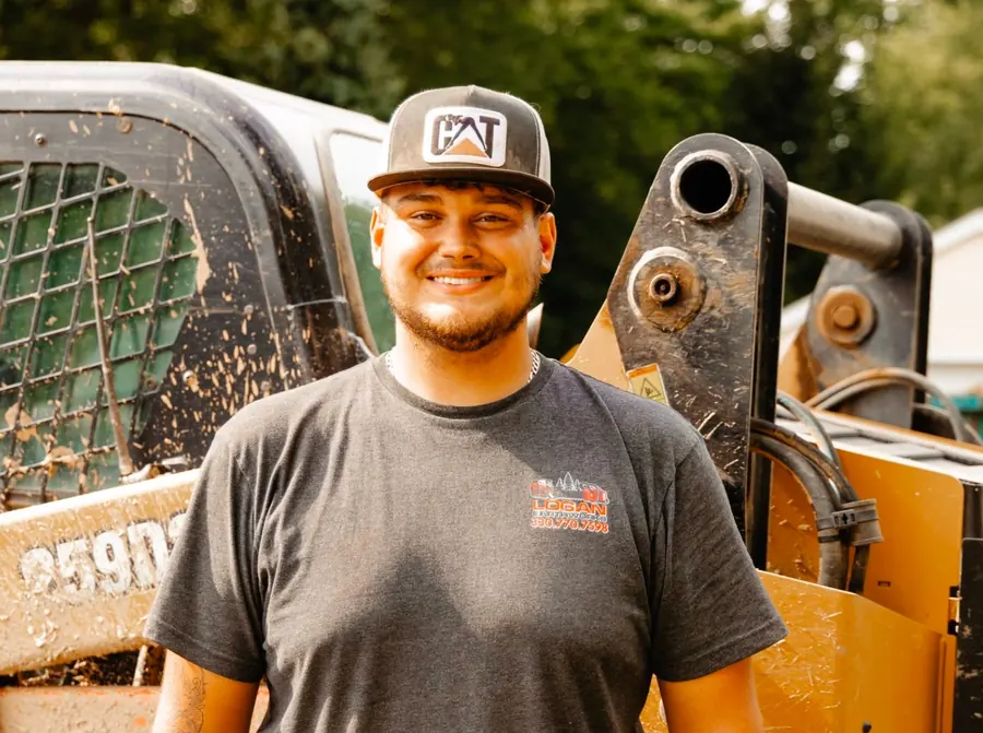 Smiling crew member in a CAT cap and Logan Earthworks shirt beside a loader