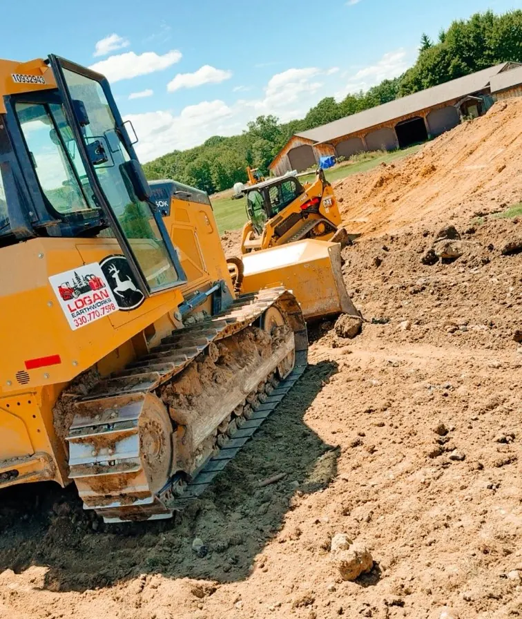 Dozer with a Logan Earthworks decal and a track loader moving dirt near a barn