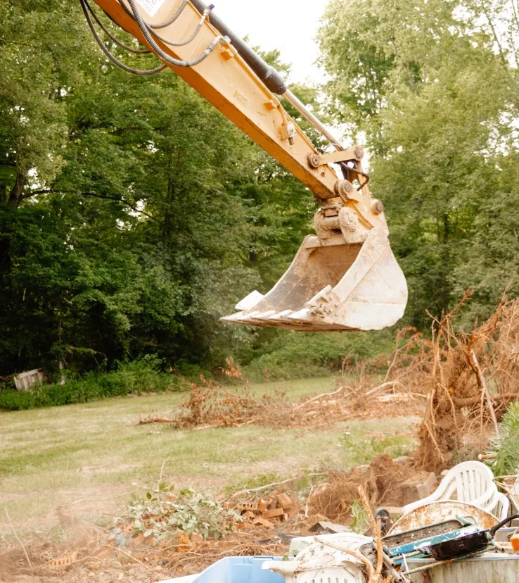 Excavator bucket raised over old lawn chairs, bricks and yard debris