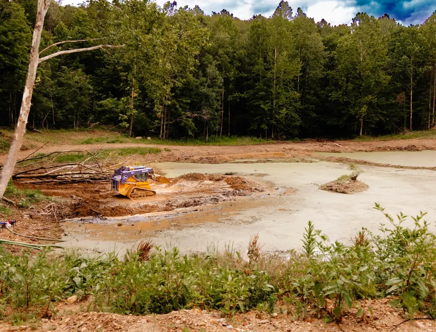 Compact track loader shaping the muddy edge of a pond ringed by woods